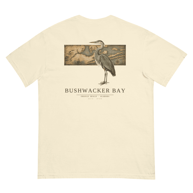 A garment-dyed tee with a back print of a great blue heron standing over an antique nautical chart of Orange Beach, Alabama, with the letterspaced wordmark Bushwacker Bay below